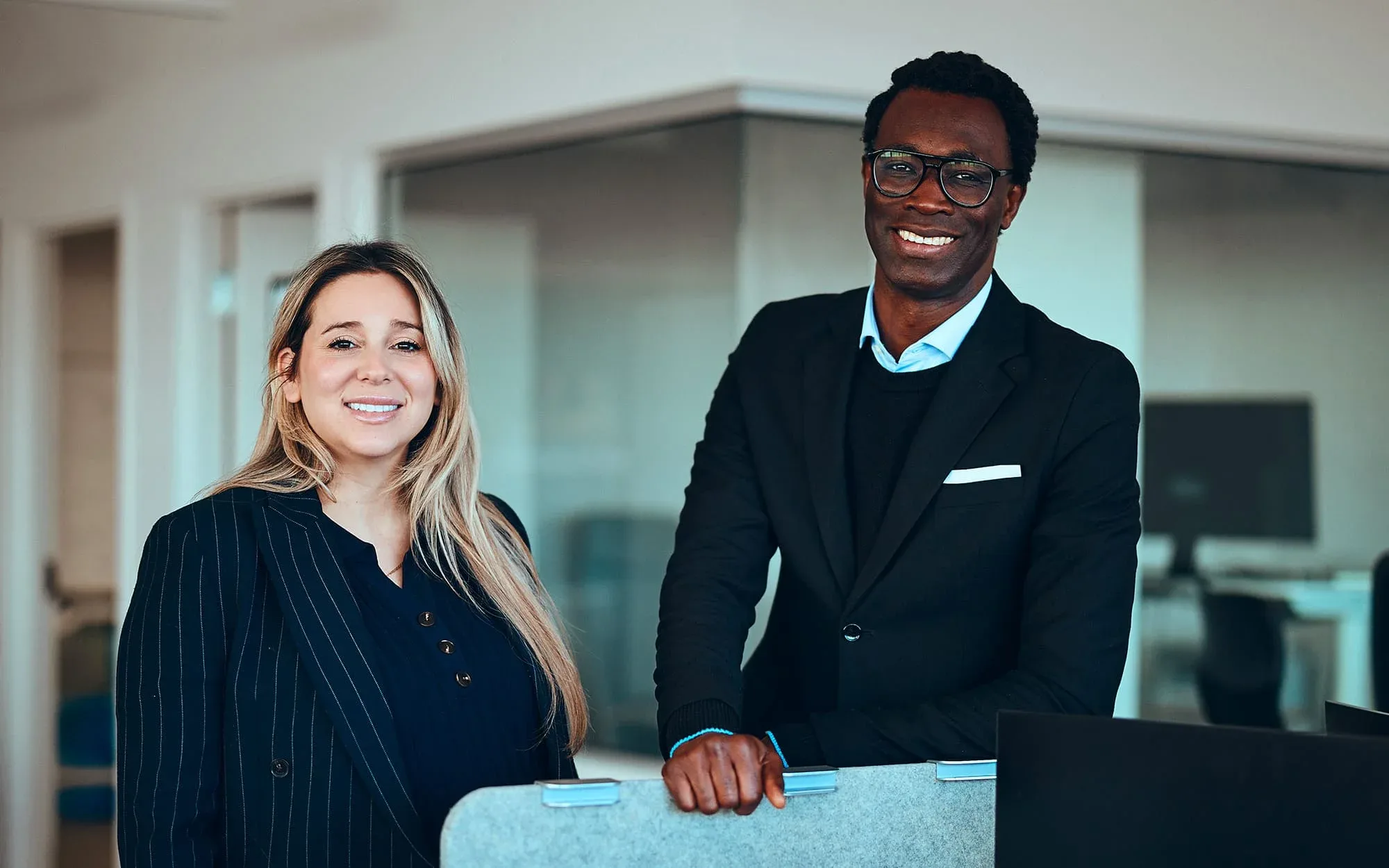 Une femme et un homme habillés chic qui regardent la caméra en souriant dans des bureaux
