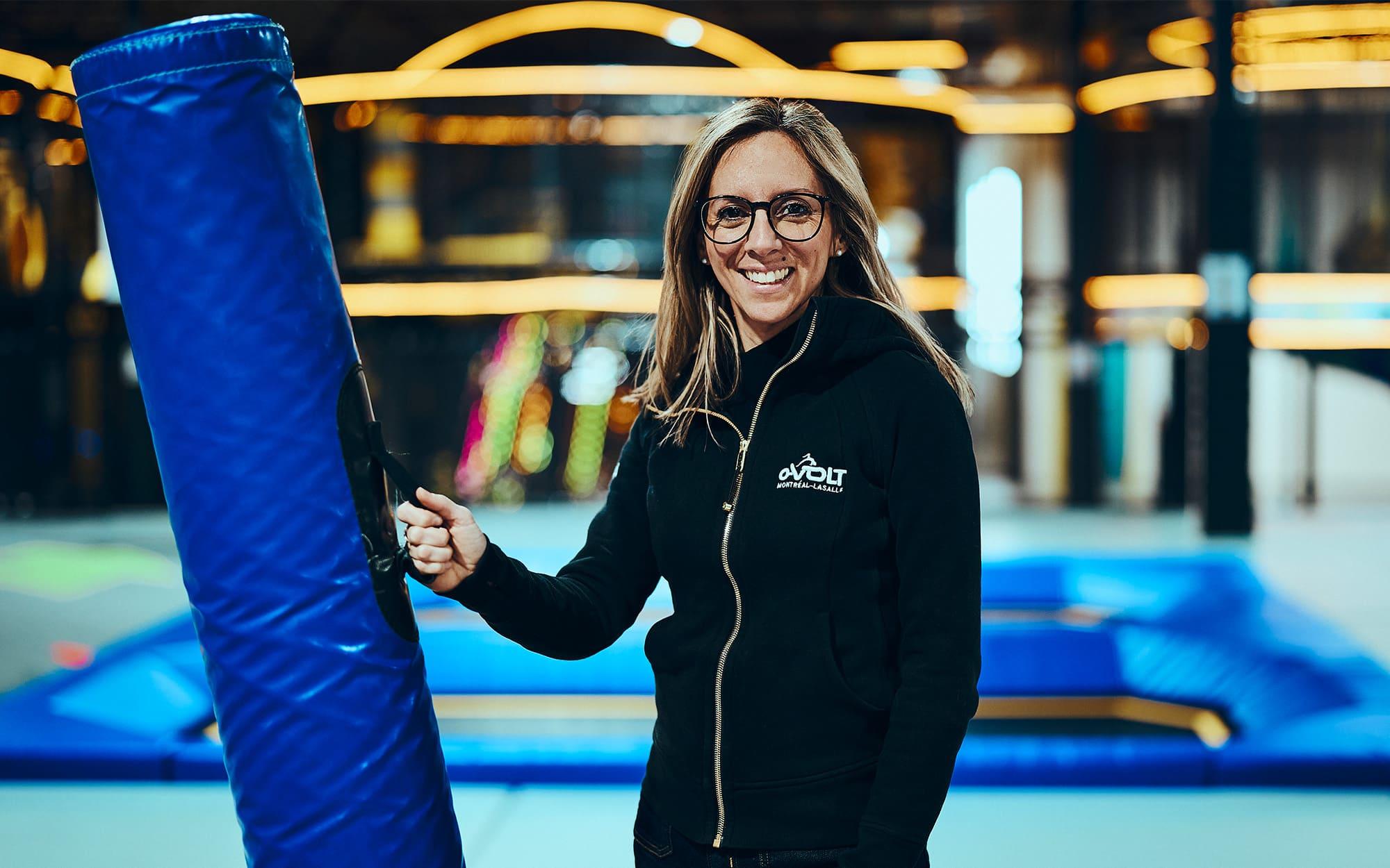 A woman inside a building in front of some trampolines, smiling at the camera while holding a protective pad