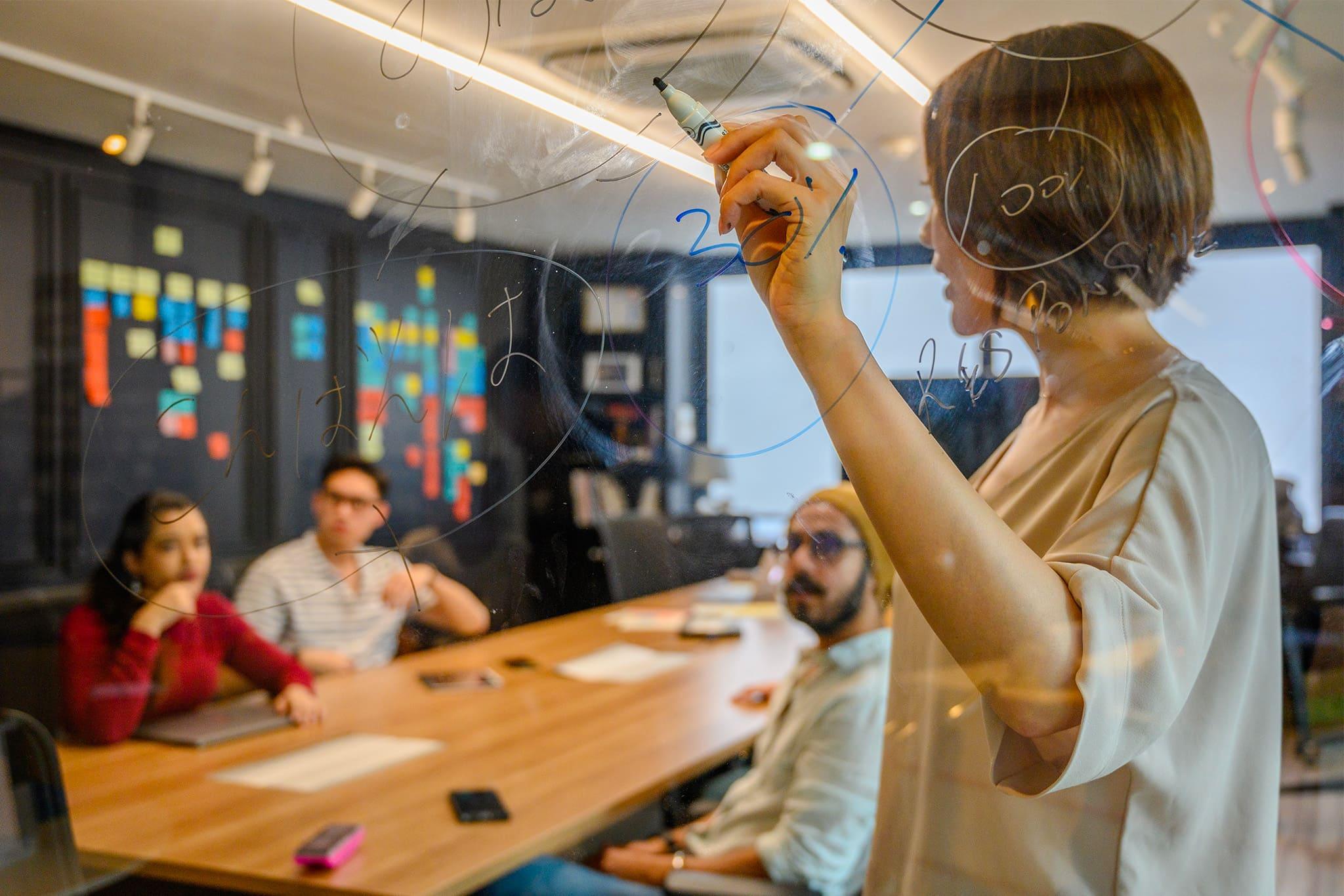 Woman standing and writing with a marker on a window. She is discussing numbers and strategy with her colleagues, who are seated at a table.